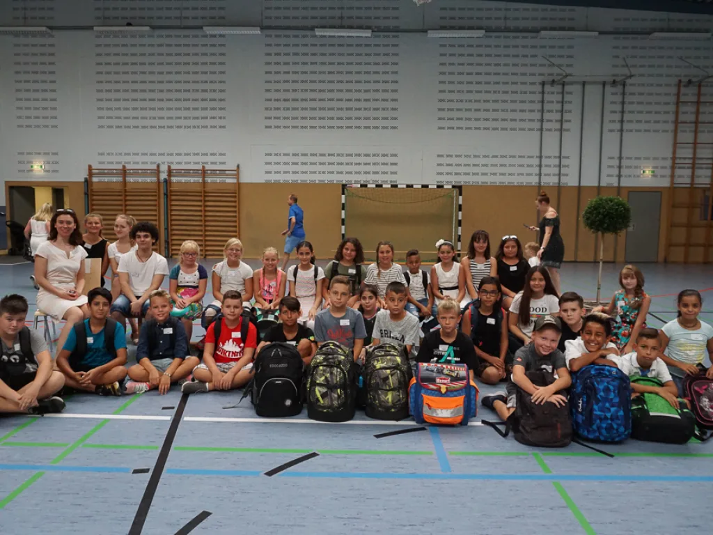Neue fünfte Klasse sitzt mit Ranzen in der Sporthalle.